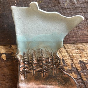 May include: A ceramic dish shaped like the state of Minnesota with a blue and white glaze on top and a brown glaze on the bottom. The dish has a design of pine trees in the brown glaze.