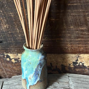 May include: A small ceramic vase with a textured, sandy-colored base and a glossy, dripping glaze in shades of turquoise, blue, and light green. The vase is filled with a bundle of thin, light brown wooden incense sticks standing upright.