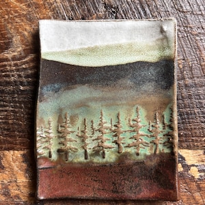 May include: A rectangular ceramic tray featuring a landscape design with a white sky, a dark brown mountain range, and a light green forest of embossed pine trees above a textured reddish-brown foreground with a smooth, glazed finish.