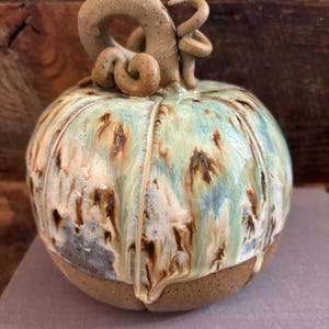 May include: A ceramic pumpkin sculpture featuring a glossy glaze in shades of turquoise, cream, and brown that drips down toward the base. The pumpkin has a textured, unglazed brown bottom and a sculpted, curled stem in a matte tan finish.