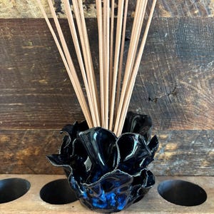 May include: A glossy, dark blue and black ceramic holder with a ruffled, organic edge, containing a bundle of thin, light brown wooden incense sticks. The vessel features a deep, reflective glaze with visible blue highlights throughout the surface.