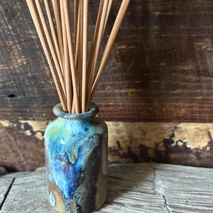 May include: A small, cylindrical ceramic vase with a reactive glaze featuring shades of deep blue, turquoise, and earthy brown. The vase holds a bundle of thin, light brown wooden incense sticks that fan out from the narrow opening at the top.