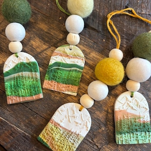 May include: Close-up of handmade decorative garlands. Each garland features ceramic tags with landscape designs, wooden beads, and felt balls in shades of green, white, and yellow. Strung on twine, these garlands are displayed on a wooden surface.