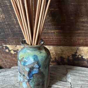 May include: A small, cylindrical ceramic vase with a mottled glaze in shades of blue, green, and brown, containing a bundle of thin, light brown wooden incense sticks. The vase has a short, narrow neck and a slightly flared rim at the top.