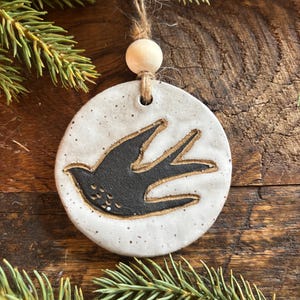 May include: A round, ceramic ornament with a black bird design. The ornament is white with speckled details and a brown outline around the bird. It hangs from a twine loop with a wooden bead. The background is a wooden surface with pine branches.
