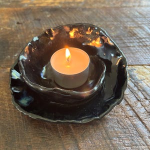 May include: A small, hand-formed black ceramic bowl with an irregular, wavy rim and a glossy finish. A lit white tea light candle with a warm orange flame sits inside the center of the bowl, casting a soft glow on the dark, reflective surface.