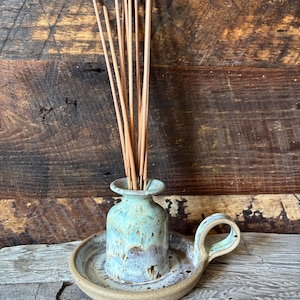 May include: A ceramic incense holder with a light blue and brown glazed finish. The holder has a small vase shape with a tray and handle. Incense sticks are placed in the vase. The holder sits on a wooden surface.