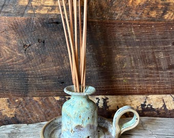 May include: A ceramic incense holder with a light blue and brown glazed finish. The holder has a small vase shape with a tray and handle. Incense sticks are placed in the vase. The holder sits on a wooden surface.