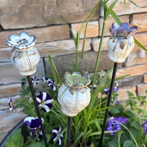 May include: Three ceramic poppy pod garden stakes in a planter with green and purple flowers. The stakes are white with brown accents and have black metal stems.