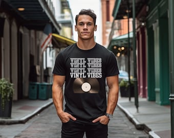 Op de afbeelding: Een zwart T-shirt met de tekst "Vinyl Vibes" die vier keer in het wit wordt herhaald. Een zwarte vinylplaat met een wit label is gecentreerd op het shirt.