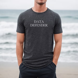 May include: Dark gray t-shirt with the text "DATA DEFENDER" in white. The shirt is a crew neck style and is made of a soft material. The person is standing on a beach with the ocean in the background.