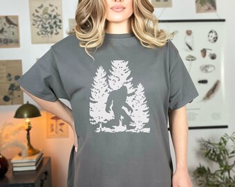 May include: Gray t-shirt featuring a white graphic of Bigfoot standing in front of two trees. The Bigfoot silhouette is detailed, and the trees are stylized. The shirt has short sleeves and a relaxed fit.