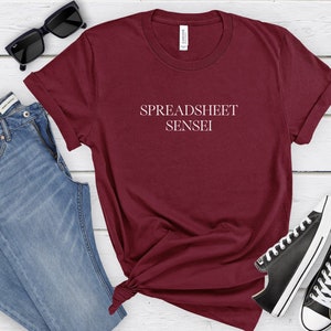 May include: A maroon t-shirt with the text "SPREADSHEET SENSEI" in white. The shirt is styled with blue jeans, black sunglasses, and black and white sneakers on a white wooden surface.