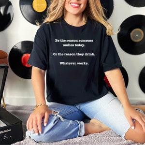 May include: A black t-shirt with white text that reads "Be the reason someone smiles today. Or the reason they drink. Whatever works."