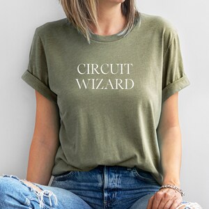 May include: An olive green t-shirt with rolled sleeves. The text "CIRCUIT WIZARD" is printed in white across the chest. The person is wearing blue jeans.