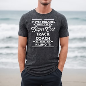 May include: A dark gray t-shirt with the text "I never dreamed I would be a Super Cool Track Coach but here I am killing it!" printed in white.