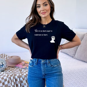 Puede incluir: Una camiseta negra con una cita blanca que dice "No hay encanto igual a la ternura del corazón." La cita está en una fuente gótica y hay una pequeña ilustración en blanco y negro de una mujer en el lado derecho de la cita.