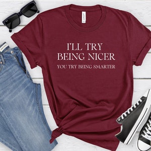 May include: Burgundy t-shirt with white text that reads "I'll try being nicer you try being smarter". The shirt is shown on a wooden surface with jeans, sunglasses, and black sneakers.