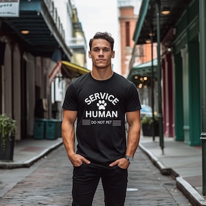 May include: Black t-shirt with white text that reads "SERVICE HUMAN" above a paw print and "DO NOT PET" below. The shirt is worn by a person standing on a cobblestone street.