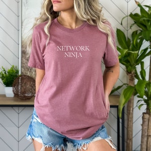 May include: A mauve-colored t-shirt with the text "NETWORK NINJA" in white block letters. The short-sleeved shirt is made of a soft material and is being worn with denim shorts. The shirt is a casual wear item.