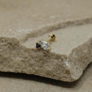 May include: A gold earring with a post and a small, round ball. It features a cluster of two gemstones: a small, square black stone and a larger, pear-shaped clear stone. The earring is designed for a piercing.