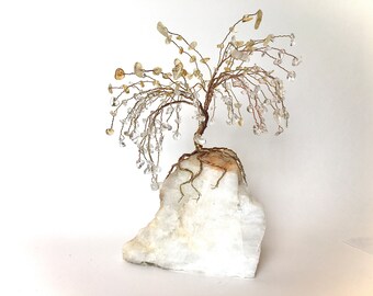 May include: A handcrafted wire tree sculpture with a brown trunk and branches adorned with clear and yellow crystal beads. The tree sits atop a large, white, rough-hewn quartz base, creating a natural and elegant decorative piece.