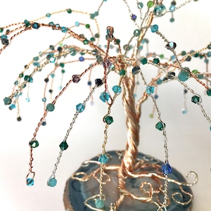 Könnte beinhalten: Eine Drahtskulptur eines Baumes mit einem Kupferstamm und Ästen, die mit blauen, grünen und violetten Kristallen verziert sind. Der Baum wird auf einer Agatscheibe präsentiert.