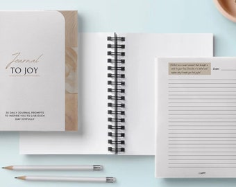 May include: A white spiral-bound journal with the text "Journal to Joy" and "30 Daily Journal Prompts". The journal is open to a lined page with a prompt. Two pencils are also visible.