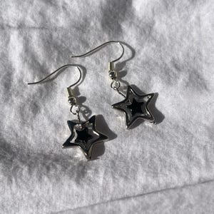 May include: A pair of silver-toned star-shaped earrings. Each earring features a layered design with a black center, creating a three-dimensional effect. The earrings have a hook closure and are displayed on a white, textured surface.