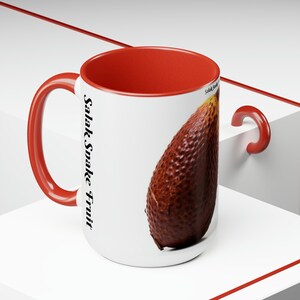 Może przedstawiać: Biały kubek ceramiczny z czerwonym wnętrzem i uchwytem. Kubek przedstawia brązowy owoc węża z nadrukiem "Salak Snake Fruit" na boku.