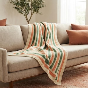 May include: A rectangular crochet blanket featuring a repeating vertical striped pattern in cream, sage green, and muted orange. The textile has a textured, woven appearance with a consistent stitch throughout, draped over a neutral-toned sofa.