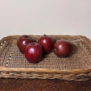May include: A rectangular woven wicker tray with a light brown finish containing four artificial dark red apples. The tray features a braided rim and a flat, grid-patterned base. The apples are smooth, round, and have small dark stems.