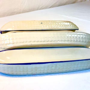 May include: A set of three elongated ceramic serving dishes shaped like corn cobs. Each dish features a cream-colored exterior with a textured kernel pattern and a smooth interior, finished with a distinct dark blue painted rim along the edges.