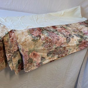 May include: A bedspread featuring a floral pattern with shades of pink, mauve, and green on a tan background. The fabric has a folded section revealing a solid cream-colored underside. The floral print includes detailed depictions of roses.