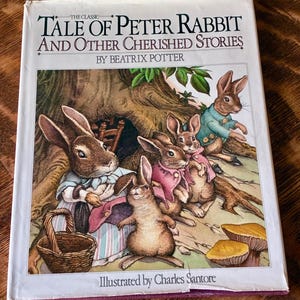 May include: A hardcover book titled The Classic Tale of Peter Rabbit and Other Cherished Stories by Beatrix Potter, illustrated by Charles Santore. The cover features a color illustration of four rabbits in clothing near a tree and mushrooms.