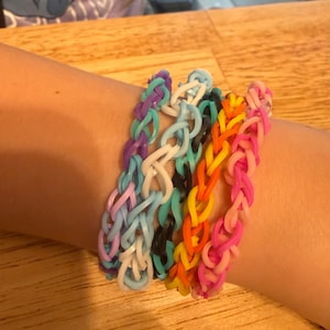 May include: A selection of colourful rubber band bracelets in shades of purple, blue, white, black, orange, yellow, and pink. The bracelets are made of woven rubber bands and are worn on a wrist.