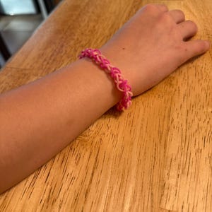 Puede incluir: Una pulsera de goma rosa y rosa claro se lleva en un brazo. La pulsera está hecha de bandas de goma tejidas. El brazo descansa sobre una superficie de madera.