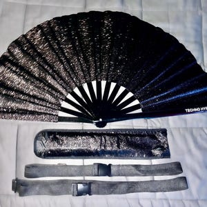 May include: A black and silver folding fan with a white and black handle. The fan has the text "TECHNO HYPE" printed on the handle. The fan comes with a black and silver carrying case and two black straps.
