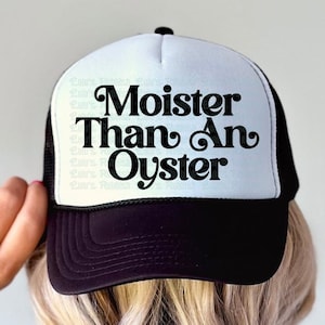 May include: A black and white trucker hat with the phrase "Moister Than An Oyster" in black script. The hat has a white front panel and a black brim and mesh back. The hat is being held by a person.