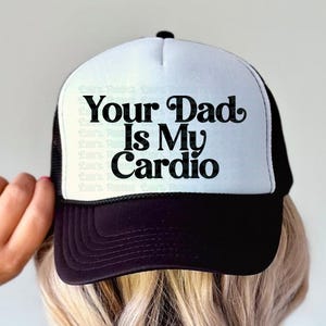 May include: A black and white trucker hat with the text "Your Dad Is My Cardio". The hat has a white front panel, a black brim, and a black mesh back. The text is in a bold, playful font.