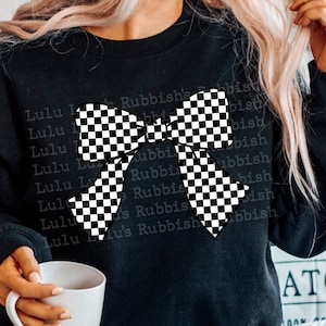 May include: Black sweatshirt featuring a large checkered bow design. The text "Lulu's Rubbish" is printed repeatedly on the shirt. The person is holding a white mug. The sweatshirt is paired with blue jeans.