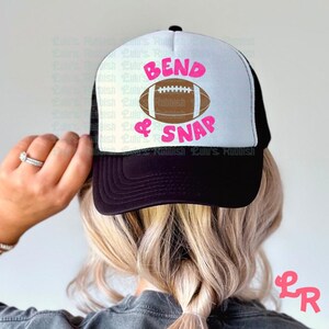 May include: A black and white trucker hat with a white mesh back. The front of the hat has a pink and white design that says "Bend & Snap" with a brown football graphic.