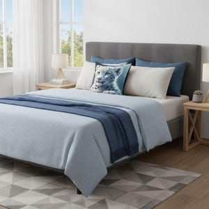 May include: A bed with a gray headboard, light blue bedding, and a navy blue throw blanket. Decorative pillows include a pillow with a snow leopard image. The bed is in a room with a geometric patterned rug and wooden side tables.