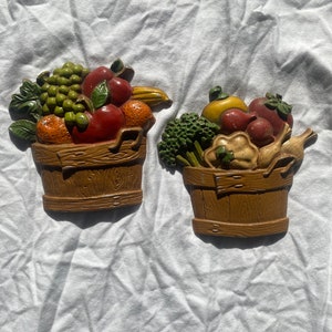 May include: Two decorative wall hangings featuring overflowing baskets of fruit and vegetables. The baskets are brown with a wood-grain texture. The produce includes grapes, apples, oranges, and broccoli, in shades of green, red, and yellow.
