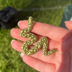 May include: A golden snake figurine with a textured finish. The snake is coiled, featuring a detailed head and body. The figurine is held in a hand against a backdrop of green grass and a white canopy.