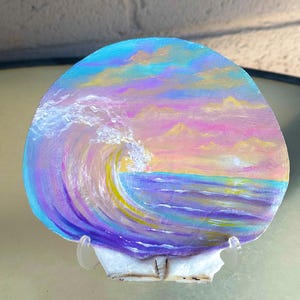 May include: A large, rounded seashell featuring a hand-painted ocean scene. The artwork depicts a curling wave in shades of purple, blue, and yellow against a sunset sky of pink, orange, and lavender. The shell is displayed on a clear stand.