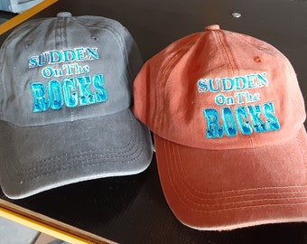 Puede incluir: Dos gorras de béisbol con el texto "Sudden On The Rocks" bordado en azul en la parte delantera. Una gorra es gris y la otra es naranja.