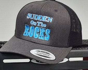 Puede incluir: Una gorra de camionero gris y negra con un logotipo bordado en azul que dice "Sudden On The Rocks". La gorra tiene una parte trasera de malla negra y una parte delantera gris con una etiqueta blanca "The Authentic Snapback Yupoong Since 1974".
