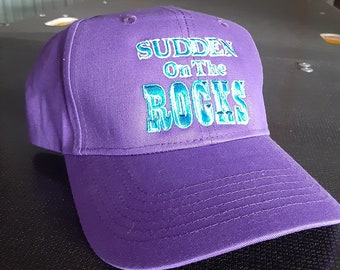 Puede incluir: Gorra de béisbol morada con el texto "Sudden On The Rocks" bordado en turquesa.