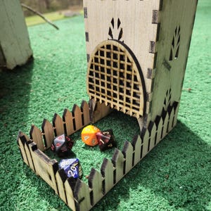May include: Wooden dice tower with a fence design and a gate opening. Four colorful dice are inside the tower. The tower is made of light brown wood and has a black and white grid pattern on the front.